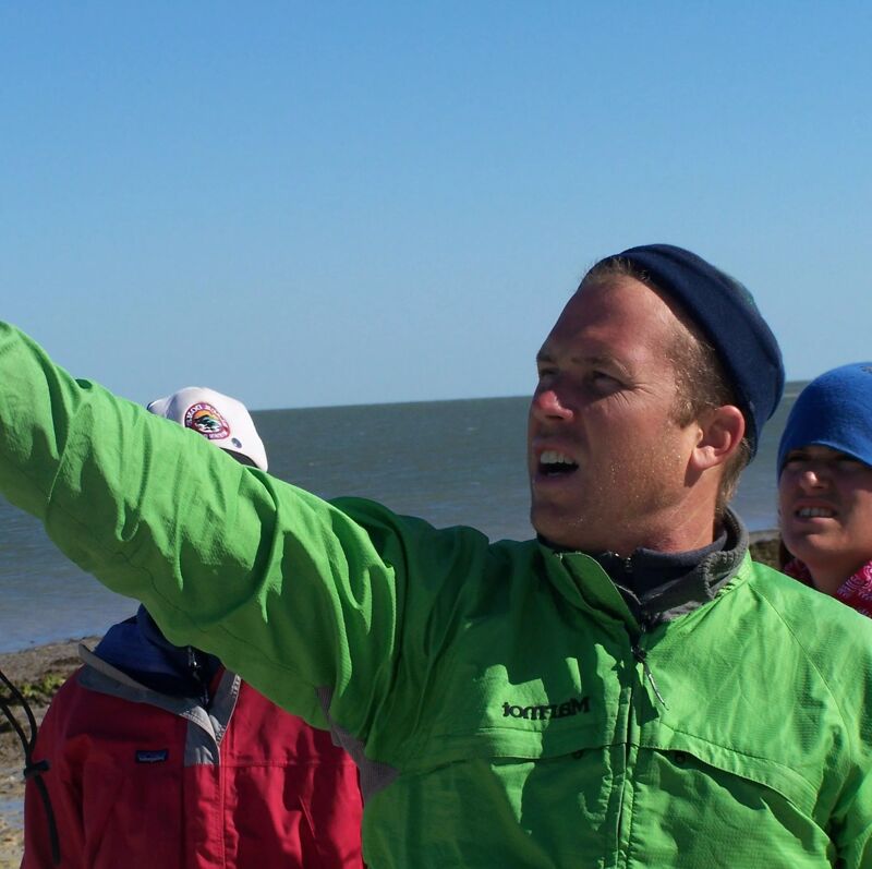 A man in a green jacket points towards the ocean. He is wearing a blue head covering. Two other people are partially visible in the background. One is wearing a white hat and a red jacket, and the other is wearing a blue hat. The sky is clear and blue, and the ocean is calm.